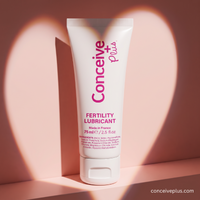 A white squeeze tube of Conceive Plus Fertility Lubricant with pink lettering, standing upright against a warm pink background with soft heart-shaped shadows.