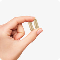 The image of a woman's left hand on a white background holding two capsules of Conceive Plus Ovulation Support in her index finger and thumb
